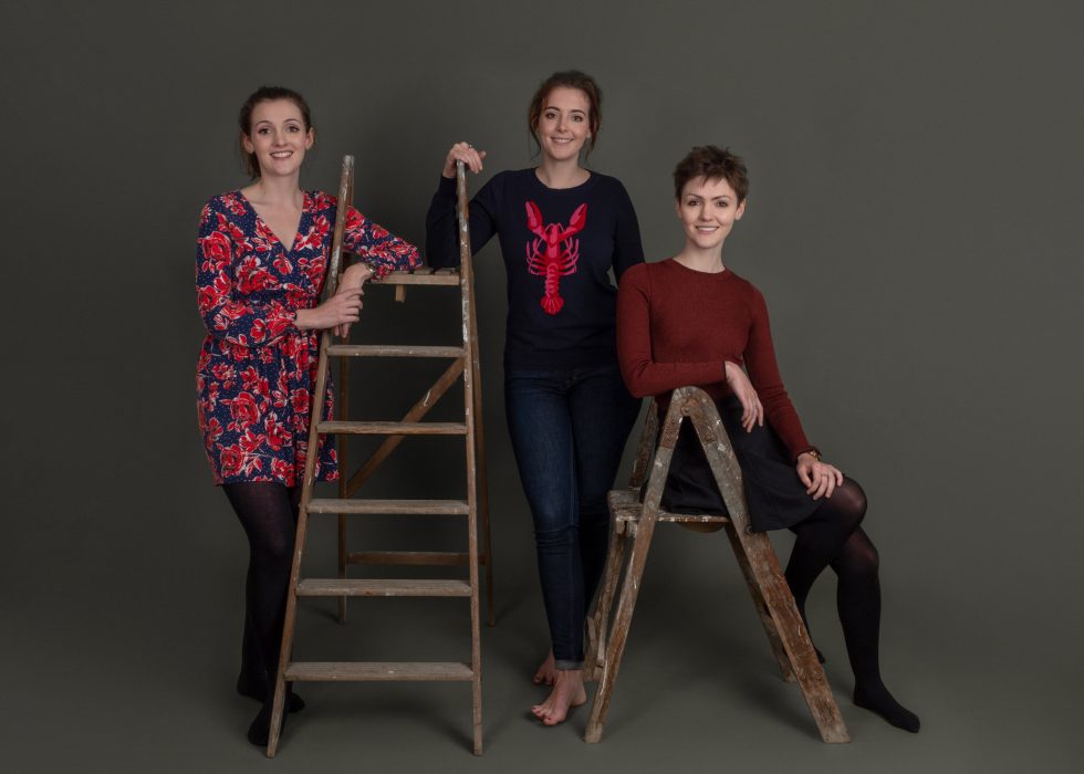 Three women posing in a studio, one standing on a wooden ladder, another leaning on it, and the third sitting on a stool, all smiling at the camera.