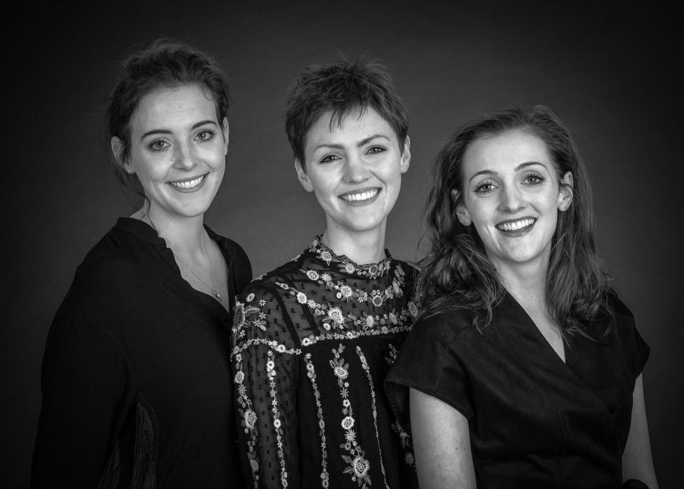 Three women smiling at the camera, with the middle one in a sequined top, flanked by two others in dark outfits, against a gray background.