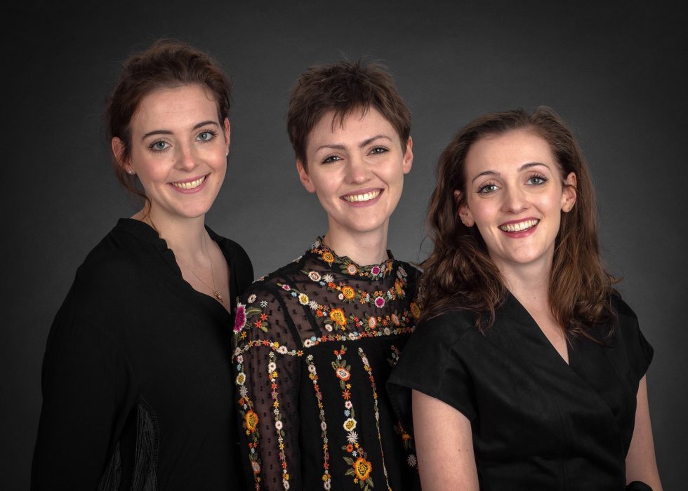 Three women smiling, with two dressed in black and one in a floral dress, standing against a dark grey background.