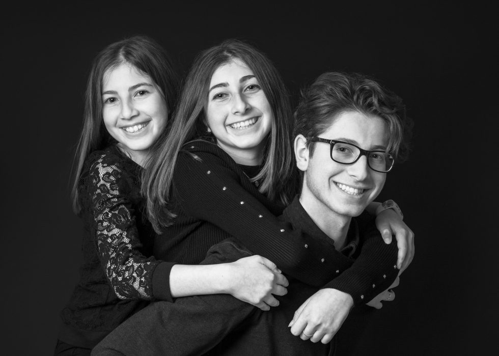 Three smiling young adults embracing each other in a joyful group hug against a dark background, in a black and white portrait.