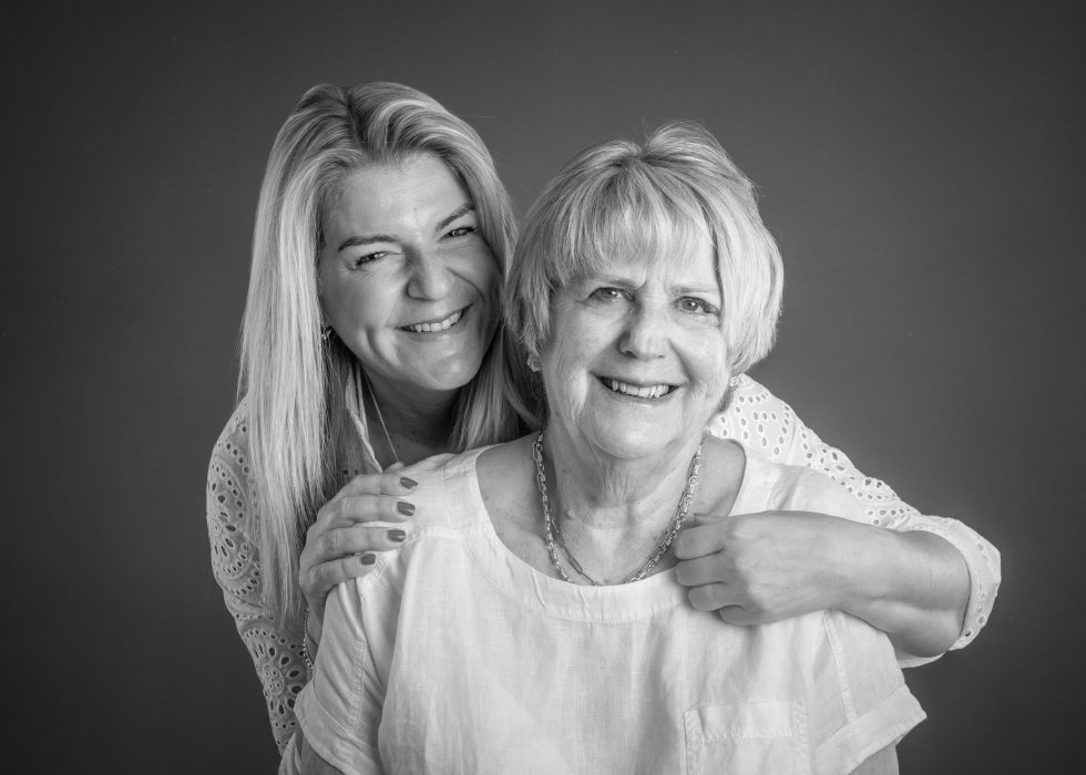 Two women smiling, with one standing behind the other and resting her hands on her shoulders, against a plain backdrop.