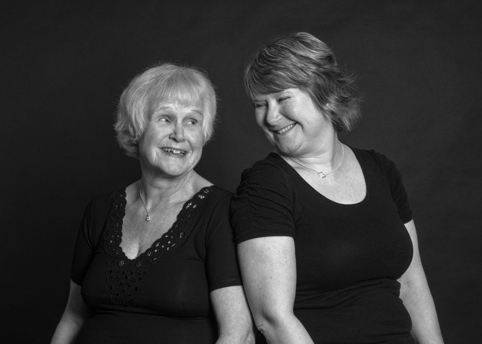 Two elderly women smiling and looking at each other, both wearing black tops, against a dark background in a black and white portrait.