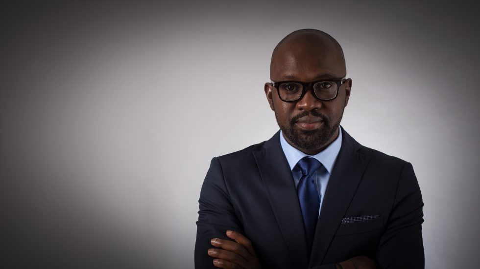 A professional portrait of a bald black man with a beard, wearing glasses and a dark suit, standing with arms crossed against a gray background.