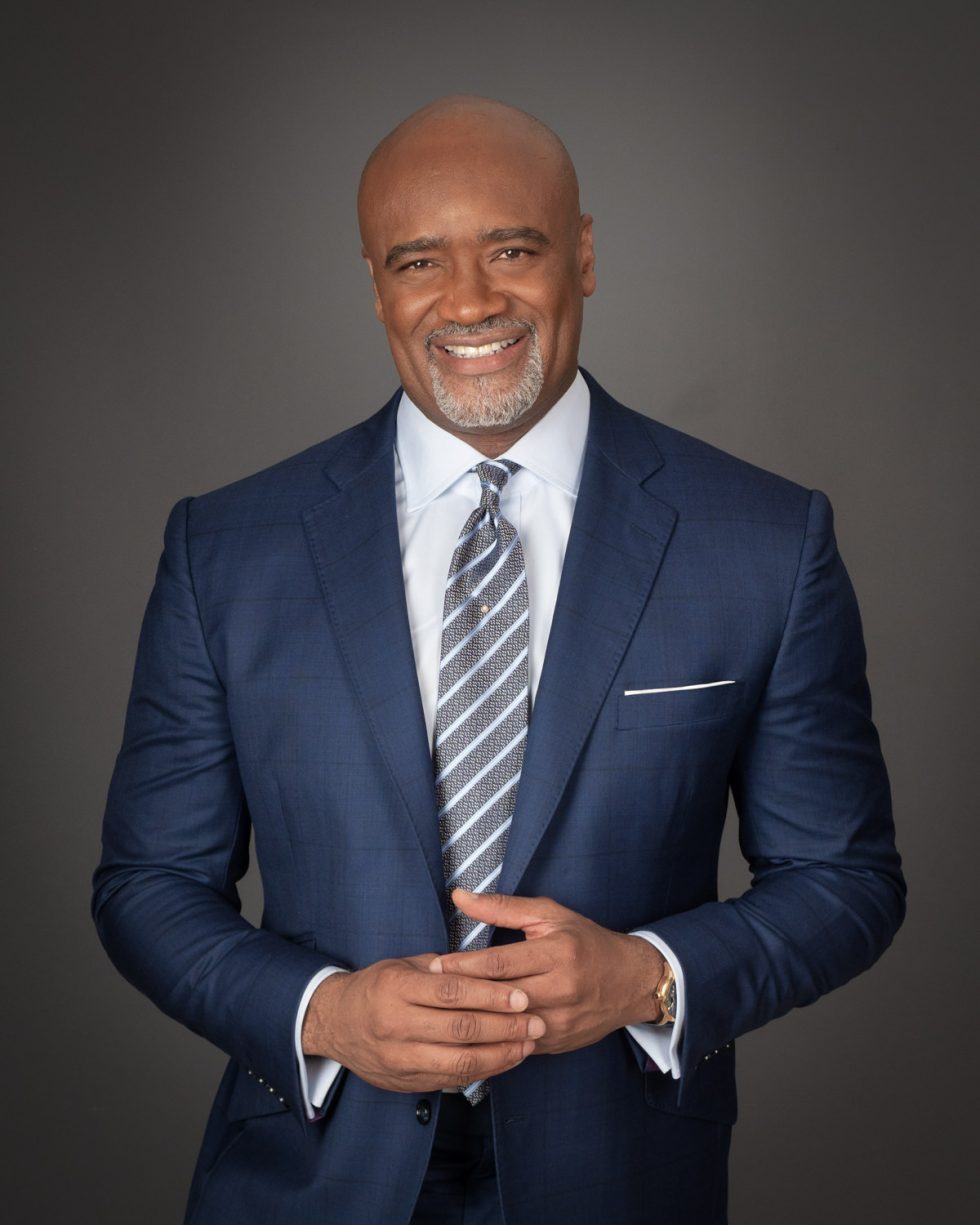 A bald man in a blue suit, white shirt, and striped tie smiling at the camera with hands clasped.