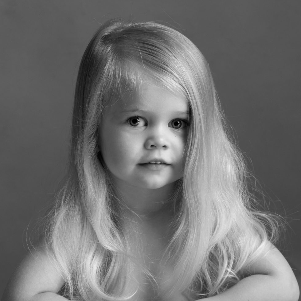 Black and white portrait of a young child with long blonde hair, looking intently at the camera.