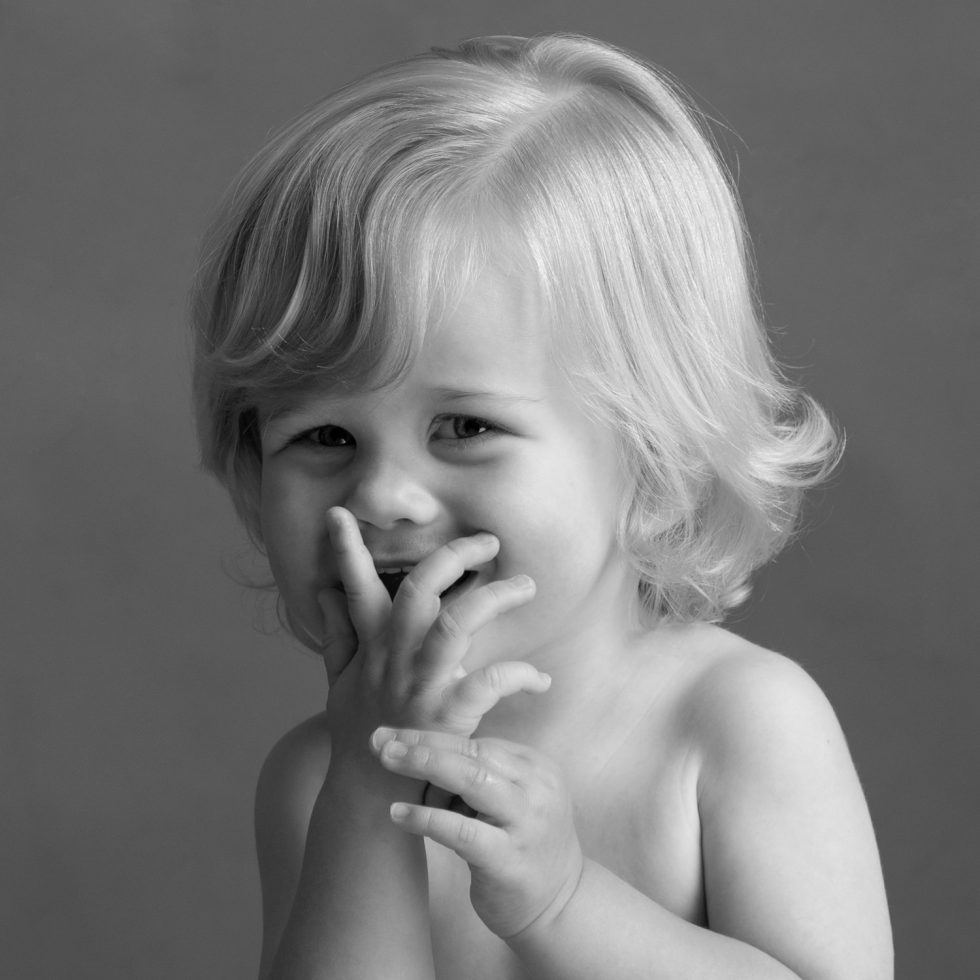A black and white portrait of a smiling toddler with curly hair, covering their mouth with their hands.