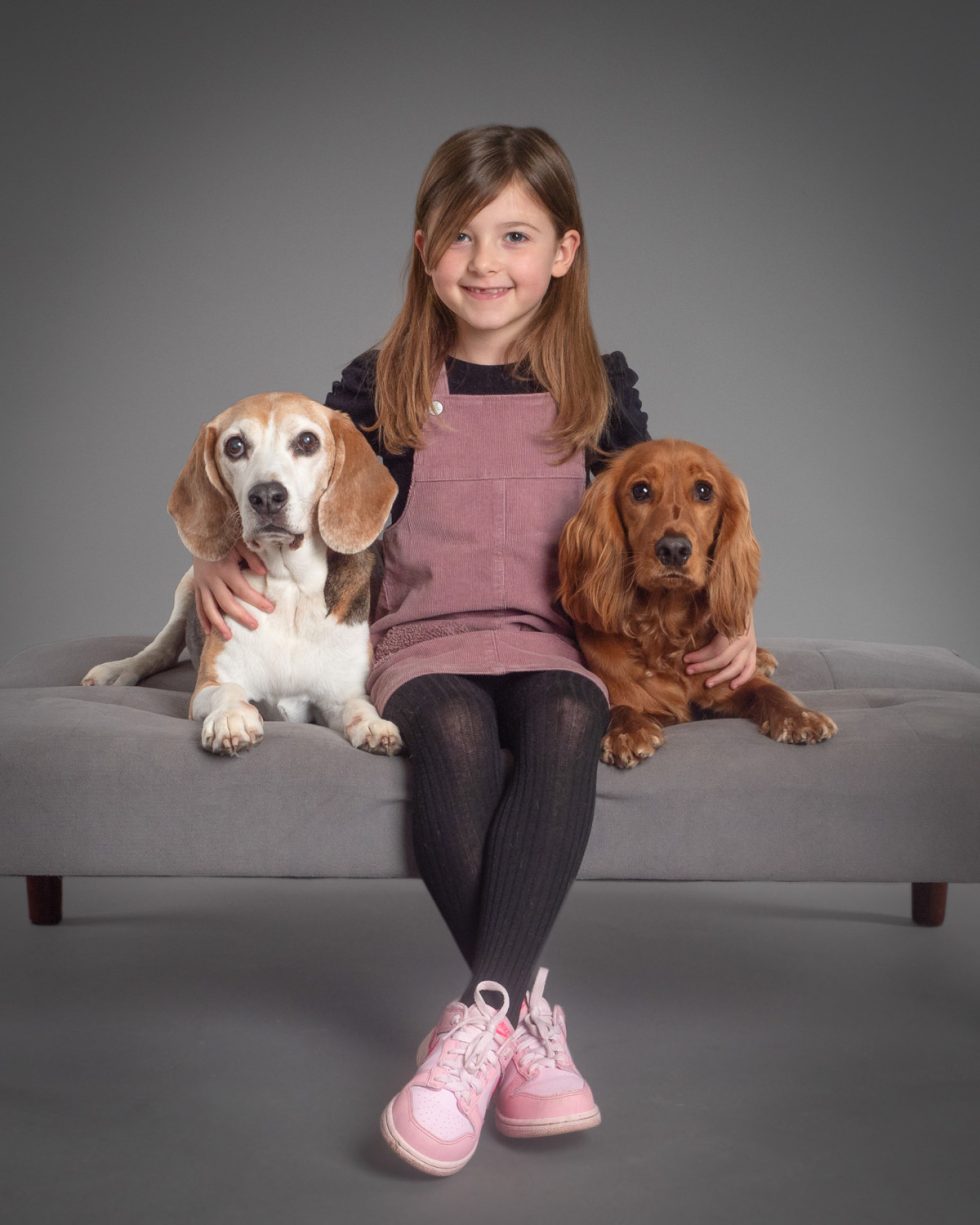A young girl in a pink dress and black tights smiles while sitting on a sofa with a beagle and a cocker spaniel, one on each side.