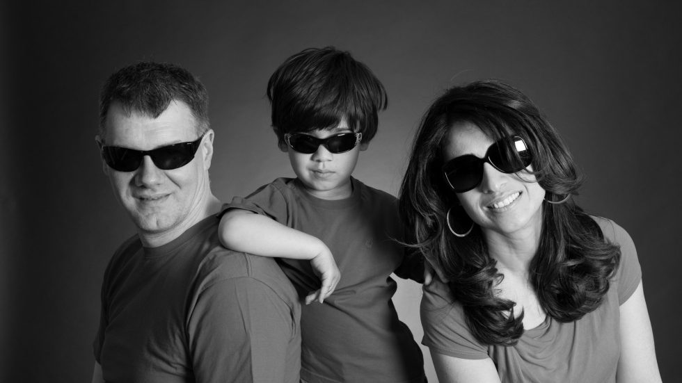 A family of three, including a man, a boy, and a woman, wearing sunglasses and posing confidently, all dressed in casual attire against a grey background.