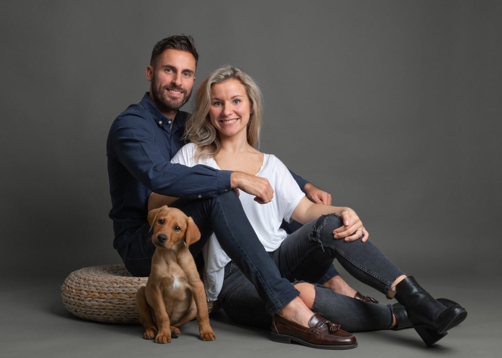 A couple sitting on a floor with a tan puppy between them, smiling at the camera, against a gray backdrop.