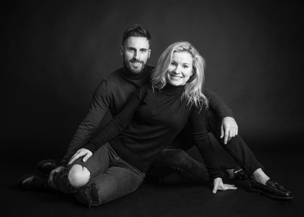 A black and white photo of a smiling man and woman sitting closely together on the ground, dressed in casual attire.
