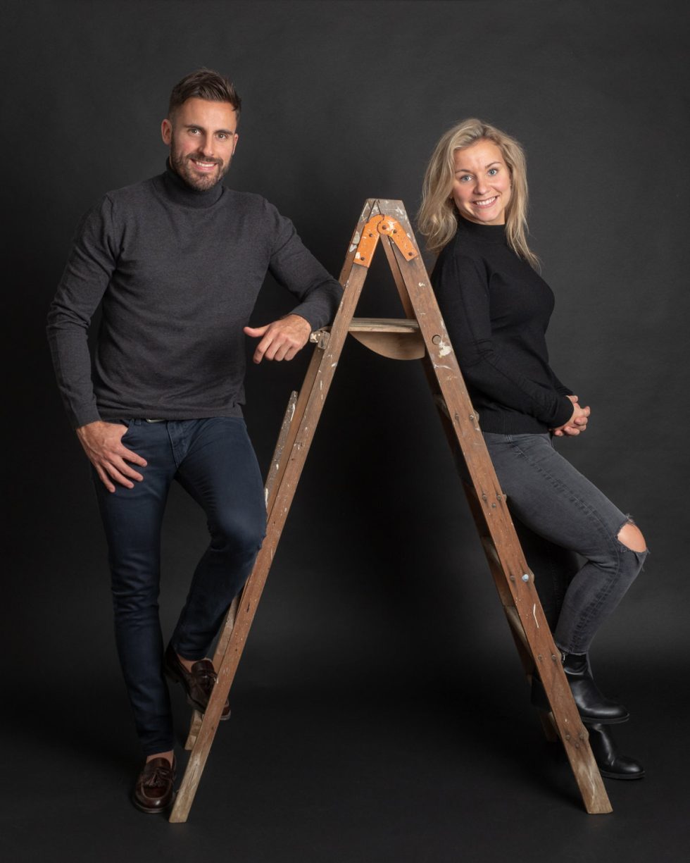 A man and a woman smiling, standing with one foot each on the rungs of a wooden ladder, against a black background.