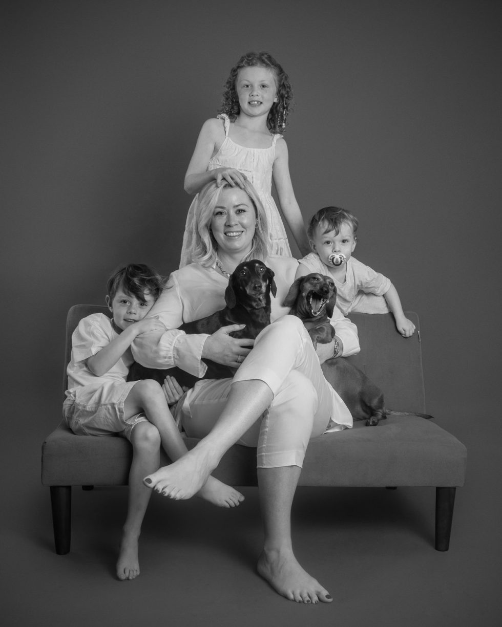 Black and white photo of a woman seated on a couch, surrounded by three young children and two dogs, all smiling at the camera.