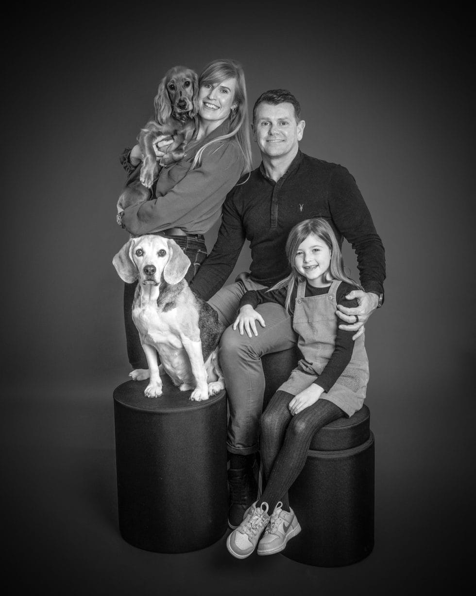 Family portrait in black and white featuring a couple, their daughter, and two beagles, posing with smiles in a studio setting.