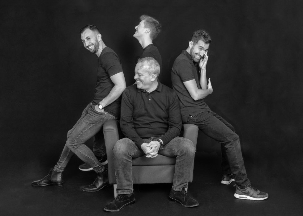 Four men in casual attire, smiling, pose in a playful line-up, each placing hands on the shoulders of the man in front, against a dark background.