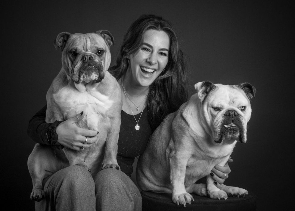 A woman smiling, sitting with two bulldogs on a dark background in a black and white photo.