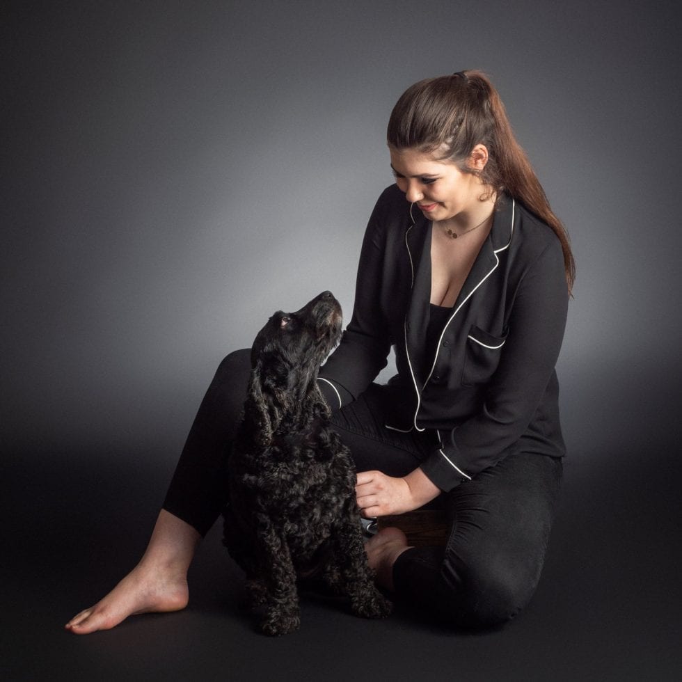A woman in dark pajamas sitting and affectionately interacting with a black, curly-furred dog against a gray background.