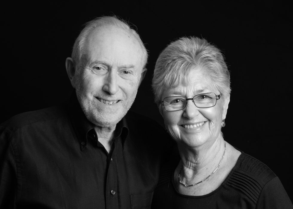 Black and white portrait of an elderly couple smiling, man wearing a black shirt and woman in a black top, against a dark background.