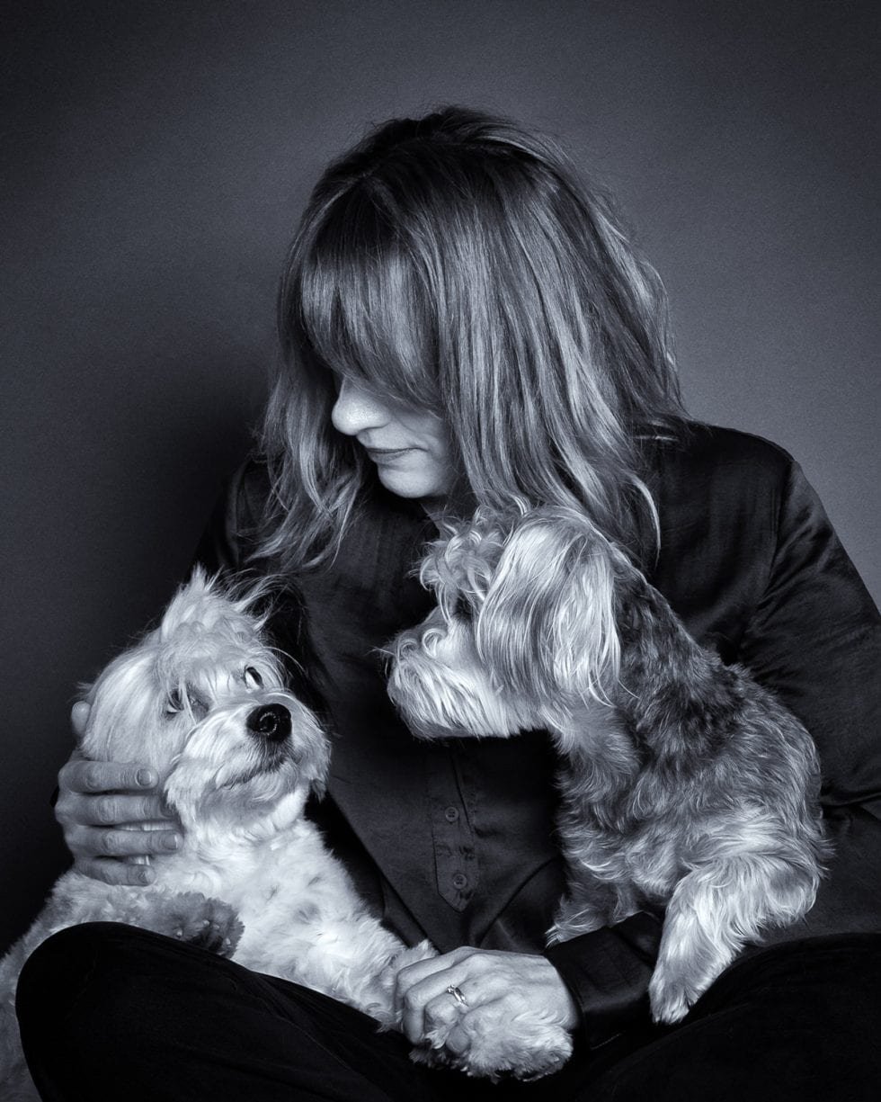 A woman in a dark shirt sits on the floor, gazing affectionately at two small dogs she's holding. the image is in black and white.