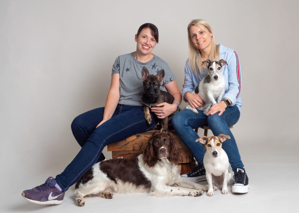 Two women smiling, seated with four dogs of various breeds on and around a wooden crate in a studio setting.