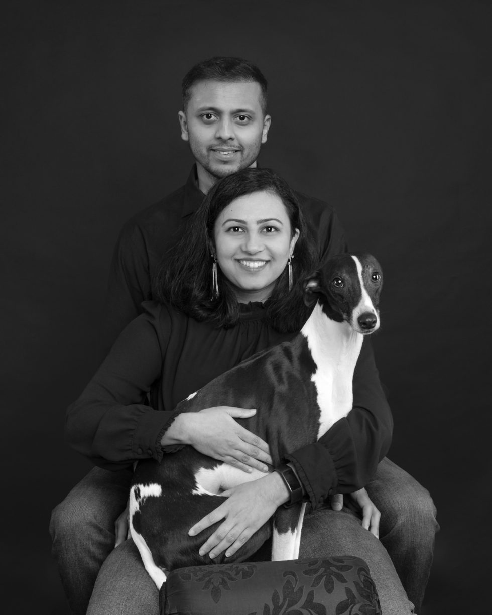 A black and white portrait of a smiling couple with a man standing behind a seated woman, both in dark clothing, holding a large black and white dog.