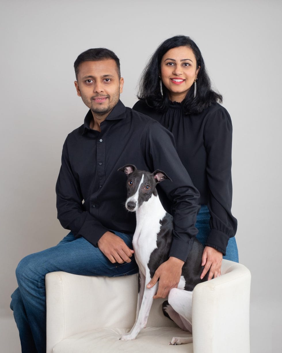 A couple and their black and white dog posing on a beige sofa, both dressed in dark clothing and smiling at the camera.