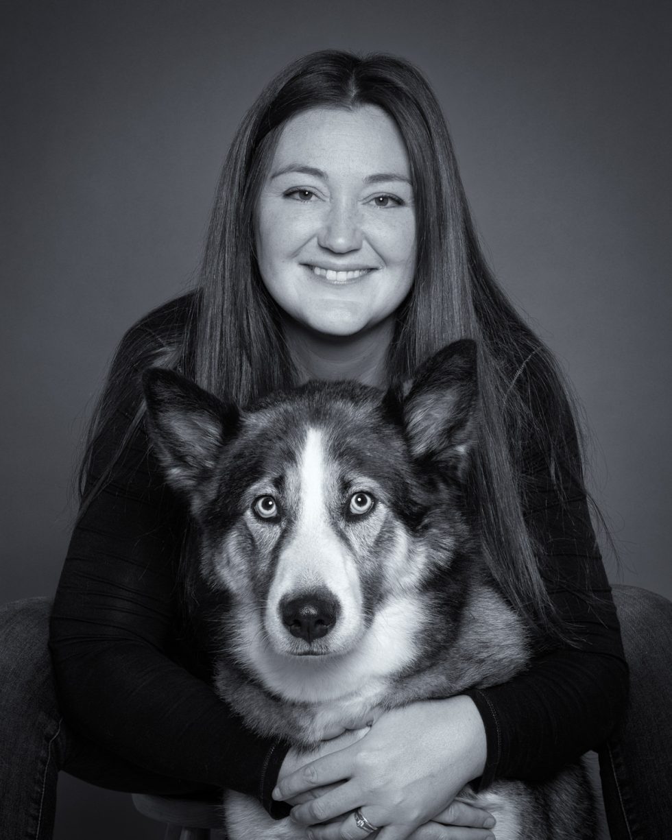 A woman smiling subtly at the camera, holding a husky with striking blue eyes. both appear in front of a simple gray background.