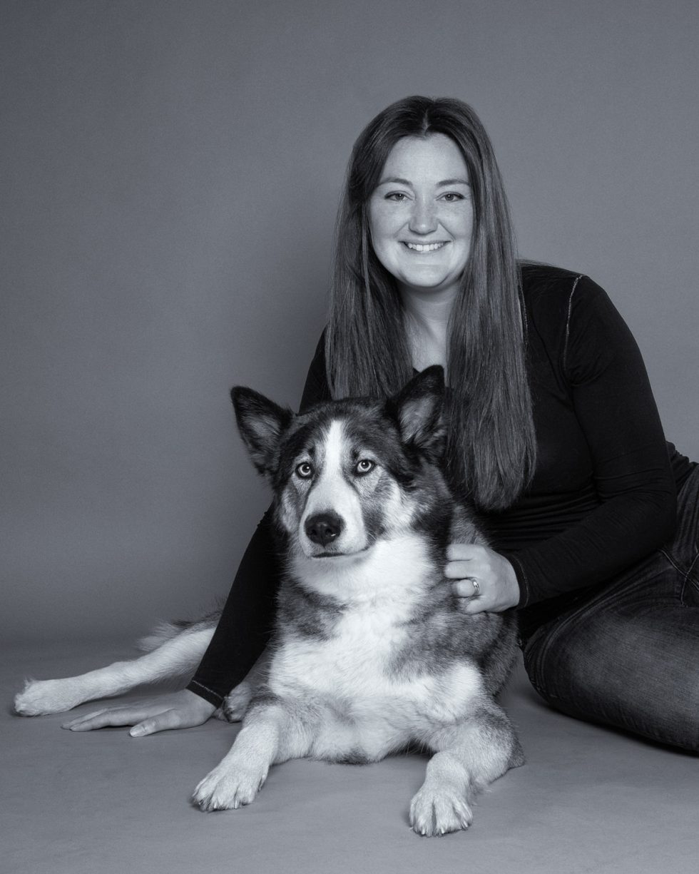 A woman smiling at the camera while sitting next to a husky with heterochromia on a gray background.