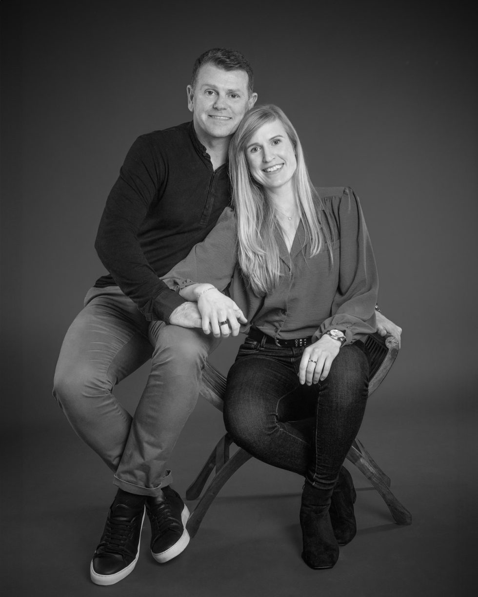 A black and white photo of a smiling man and woman seated together on a stool, both dressed in casual attire.
