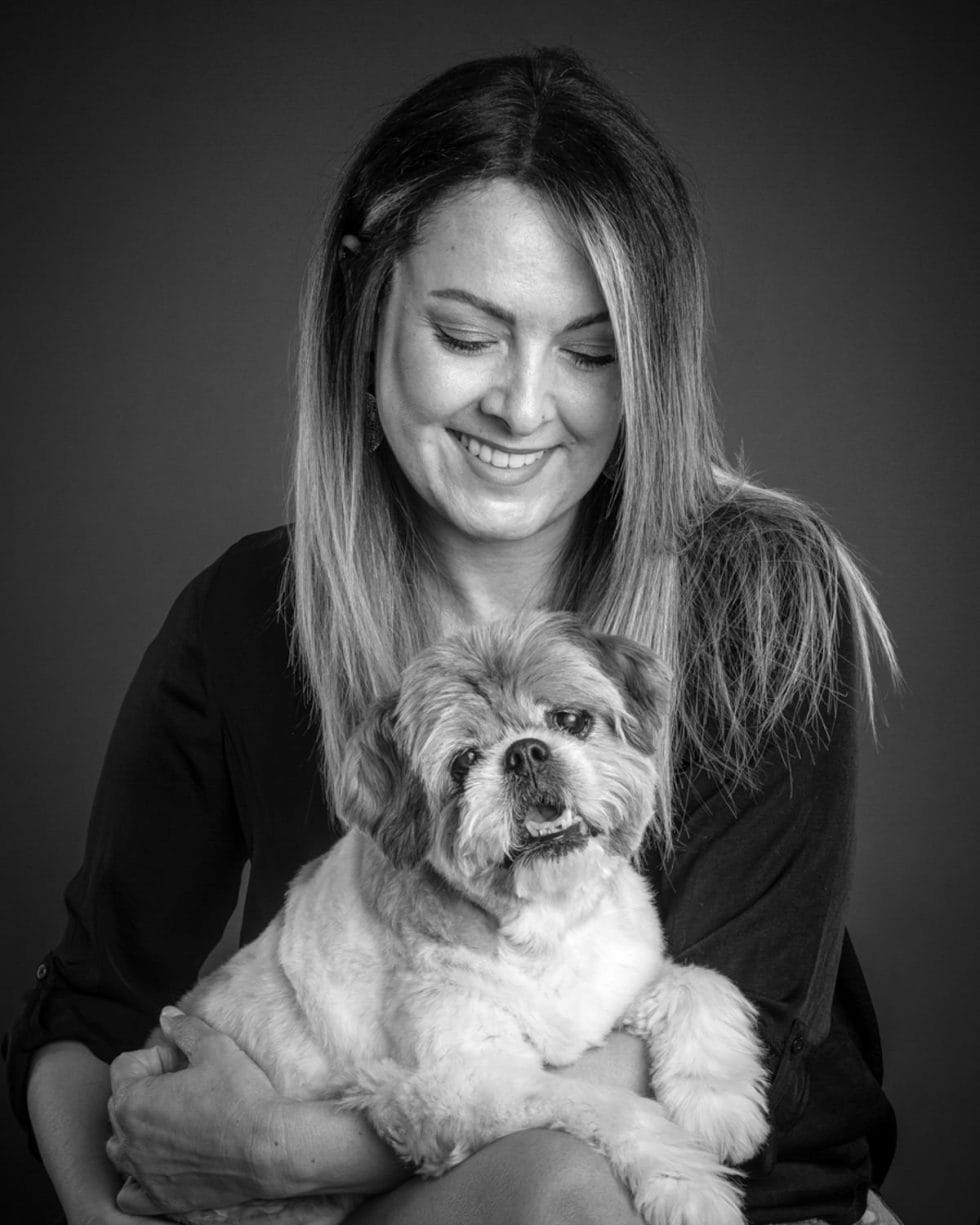 A black and white photo of a smiling woman holding a small fluffy dog in her lap.