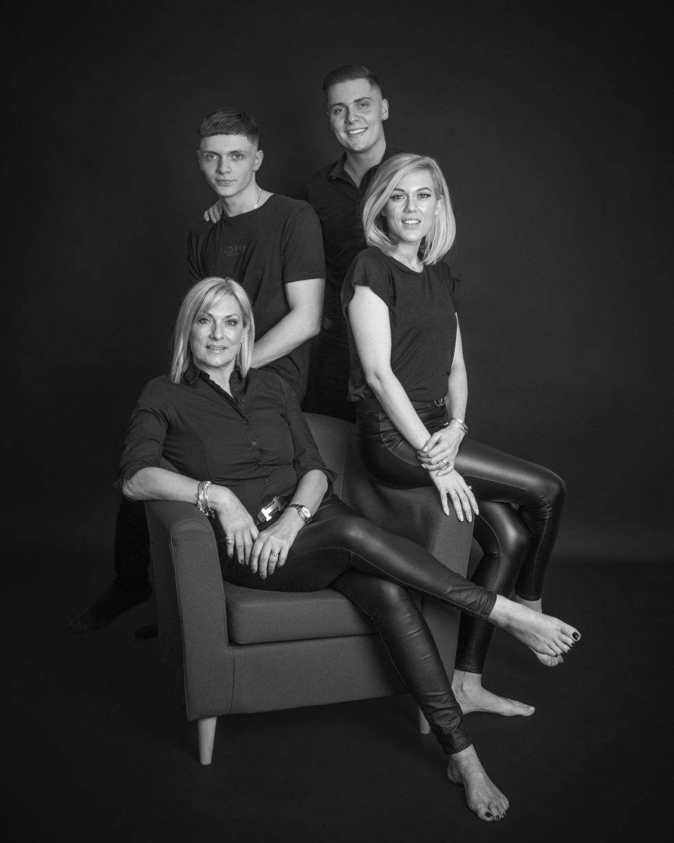 Black and white photo of a family of four, with two young men standing behind a seated woman and another woman in elegant attire, all wearing dark clothes against a black background.