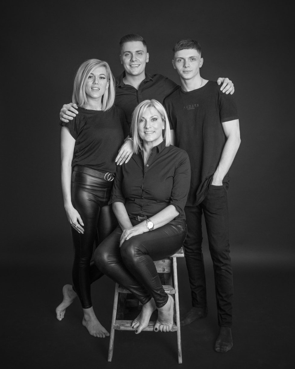 Four people in black attire posing for a studio portrait, one seated on a stool and three standing behind her, against a dark background.