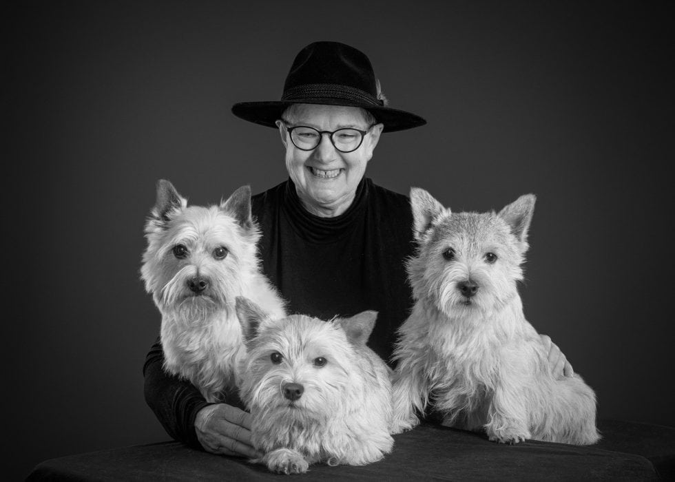 Elderly woman wearing glasses and a black hat, smiling with three west highland white terriers, in a monochrome portrait.