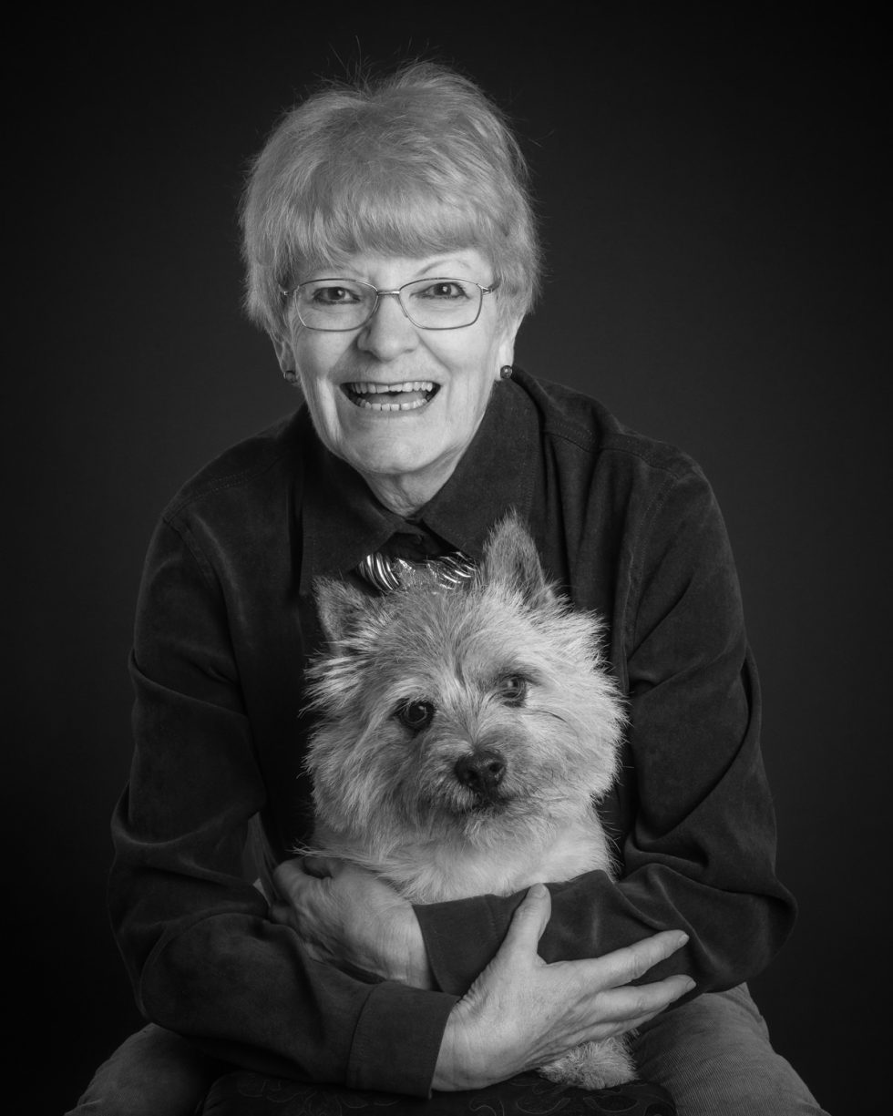 Elderly woman with glasses smiling, holding a small fluffy dog in her lap, both posing against a dark background.