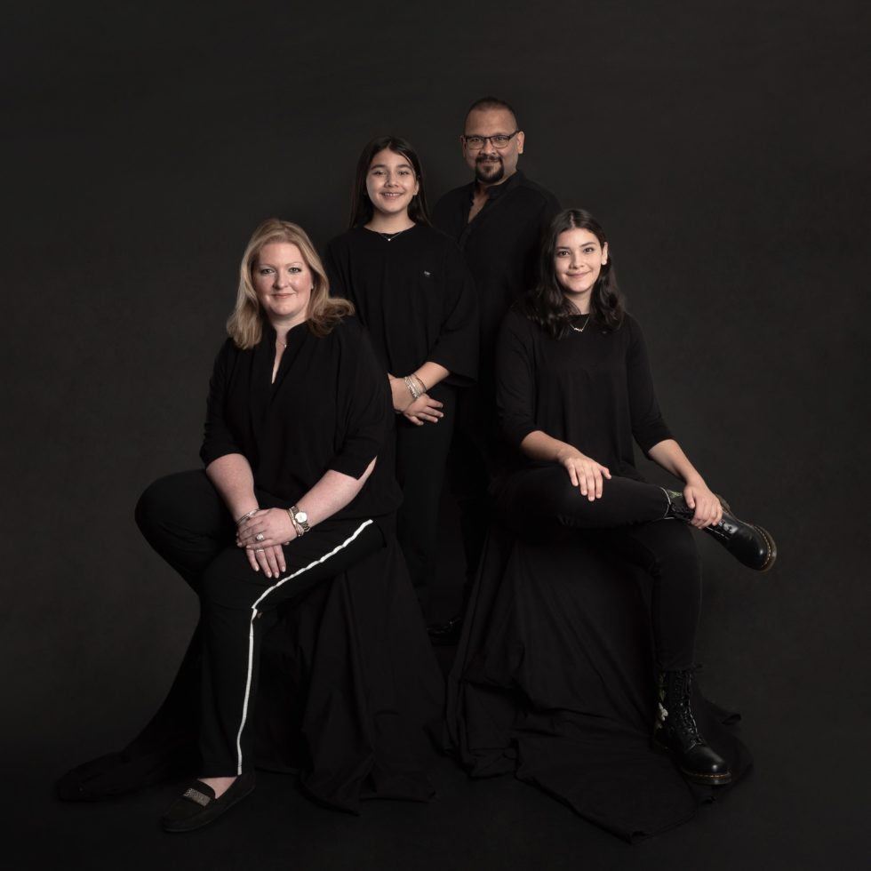 Four people dressed in black sit and stand against a dark background, looking at the camera.