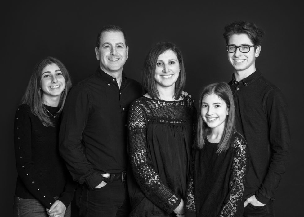 Black and white portrait of a smiling family of five, with two adults and three teenagers, dressed in dark casual clothes, standing against a dark background.
