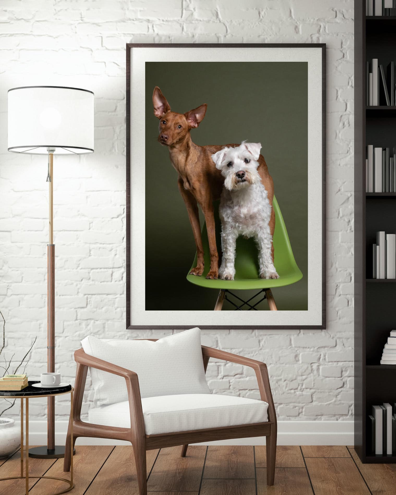 A framed picture of two dogs, one brown and one white, sitting on a green chair hangs on a white brick wall above a modern armchair and side table with a lamp.
